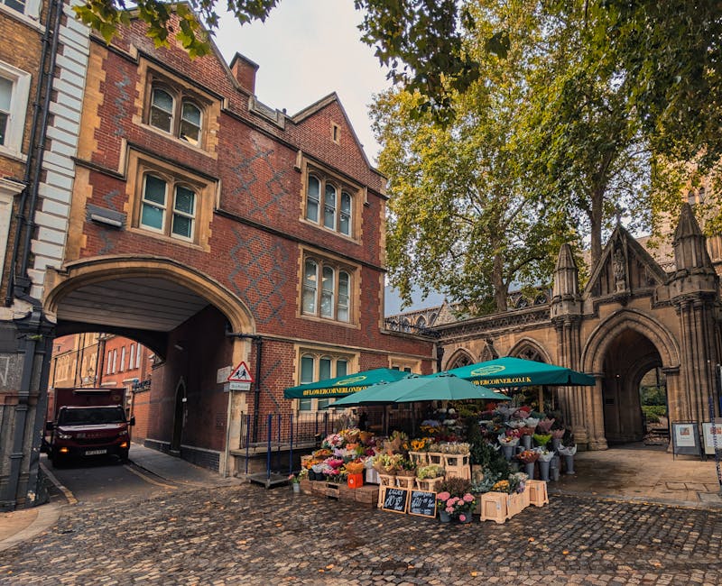 A vibrant flower stall set up on a cobblestone street in front of an historic red-brick building with arched windows and a tunnel underpass. The stall is shaded by large green umbrellas displaying the