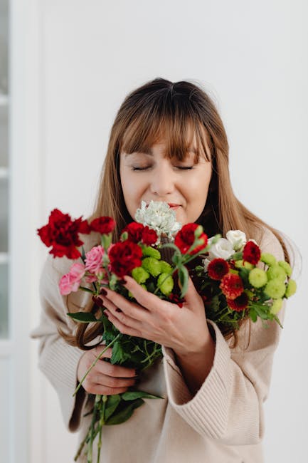 A young woman with long brown hair and closed eyes holds a bouquet of fresh flowers close to her face, expressing delight. The bouquet features vibrant red carnations, small pink roses, white and gree