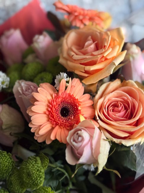 A close-up arrangement of fresh flowers including peach and pink roses with tightly curled petals, a peach gerbera daisy with a dark center, and small pink and coral blossoms. The bouquet is wrapped i