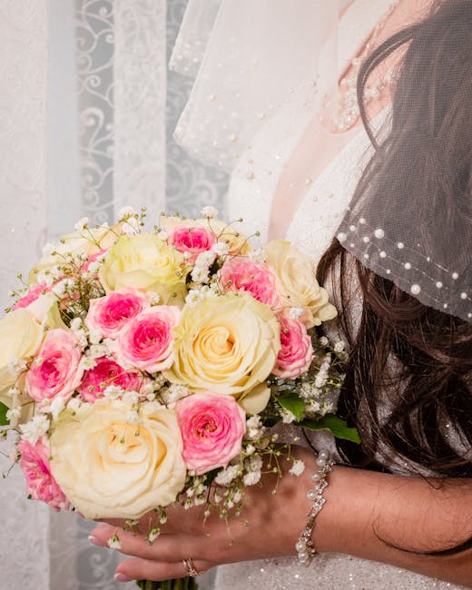 A close-up of a bride holding a floral bouquet featuring large cream-colored roses and smaller pink roses, accented with delicate baby's breath. The bouquet is arranged in a rounded, lush style with g