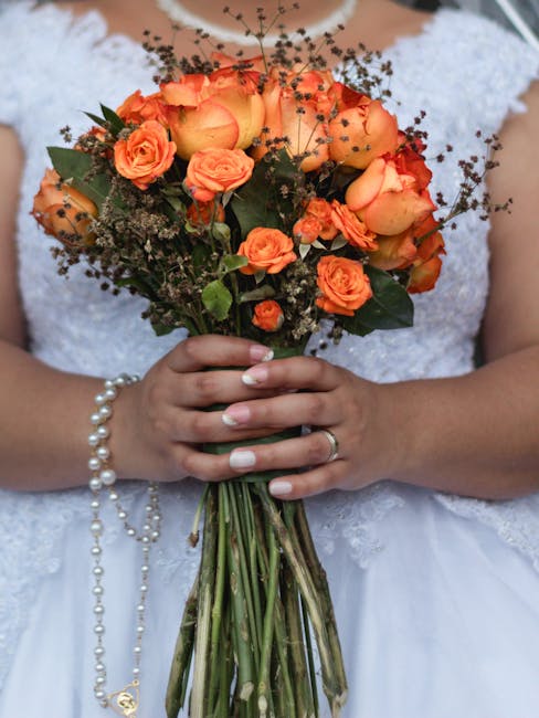 A bride holding a vibrant bouquet of fresh orange roses, peach-colored roses, and small delicate filler flowers, arranged in a loose, natural style. The bouquet features lush green foliage and is wrap