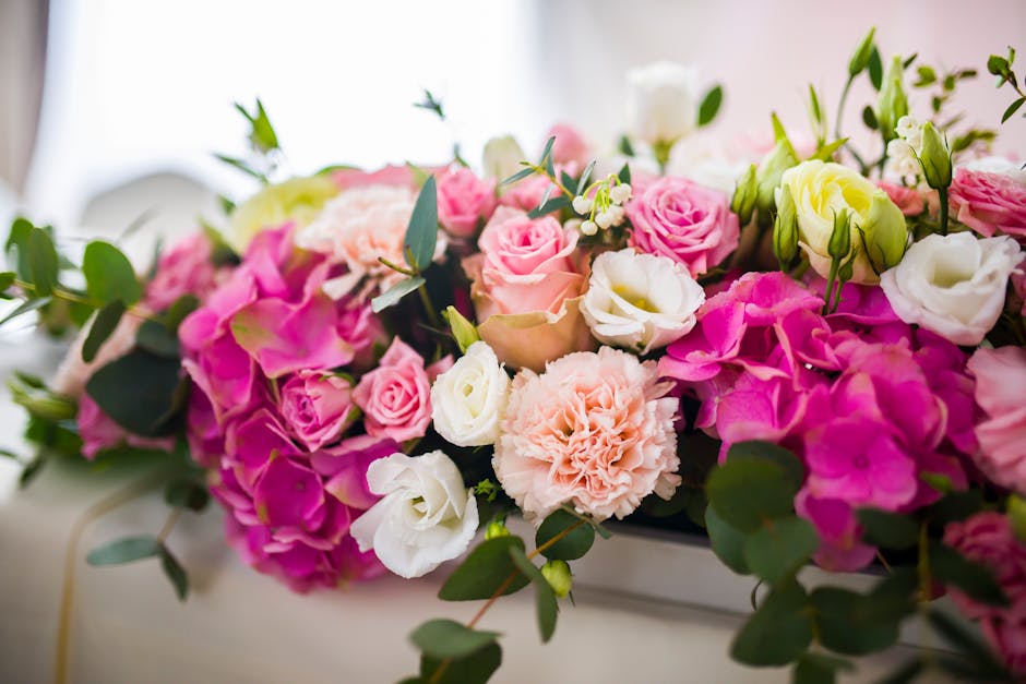 A lush, abundant floral arrangement featuring pink roses, white lisianthus, pale peach carnations, and blush pink hydrangeas, accented by dark green eucalyptus leaves. The flowers are fresh, with peta