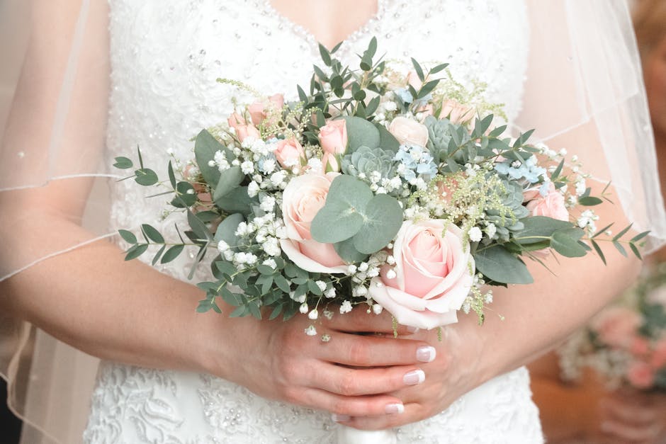A bride holding a bouquet arrangement featuring soft pink roses, white baby's breath, and eucalyptus leaves, with a mix of textured petals and fresh green foliage. The bouquet is wrapped in delicate w