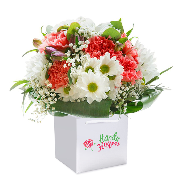 In the bustling warmth of Redbridge, a stunning bouquet comes to life within a crisp, square white gift bag-a vision of local artistry and botanical freshness. The arrangement centers around a lush medley of coral and peach carnations, every petal soft and frilled for a plush, inviting texture, interspersed with the purity of radiant white daisies, whose bright, lime-tinged centers bring vibrancy and cheer. Scattered delicately across the bouquet, clouds of white baby's breath lend a feather-light, romantic feel, while a few unopened buds with hints of purple and pink nestle in, their quiet presence hinting at transformation and renewal. Surrounding the flowers, an abundance of deep green, broad leaves frames the bouquet in luxuriant style, much like the cultivated gardens and leafy avenues found along Redbridge's streets. The sleek white gift bag, with its smooth handles, ensures easy gifting and delivery whether for birthdays, anniversaries, or just because-a trusted choice for locals. Illuminated by balanced, gentle light and set against an immaculate background, the entire composition feels fresh, abundant, and celebratory, spotlighting every thoughtful detail from blossom to foliage. It is a bouquet crafted with the care and expertise of Redbridge Florist, echoing a sense of place and offering a tangible link between loved ones across the community. Perfect for those seeking a meaningful, beautifully presented floral arrangement delivered straight to a Redbridge door.